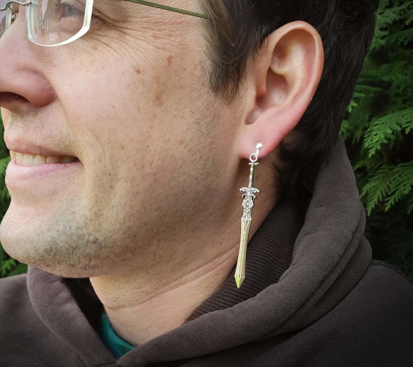 A male model wearing long silver sword earrings with clip ons. He is outside in nature and looks happy.