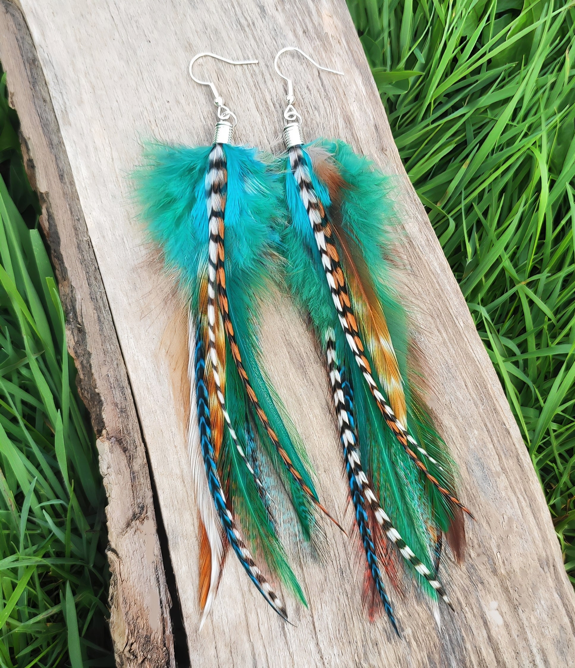 Long stripy green, white, brown, blue and black feather earrings dangling from silver stainless steel hooks. The earrings have a funky bohemian vibe, a boho hippie style.