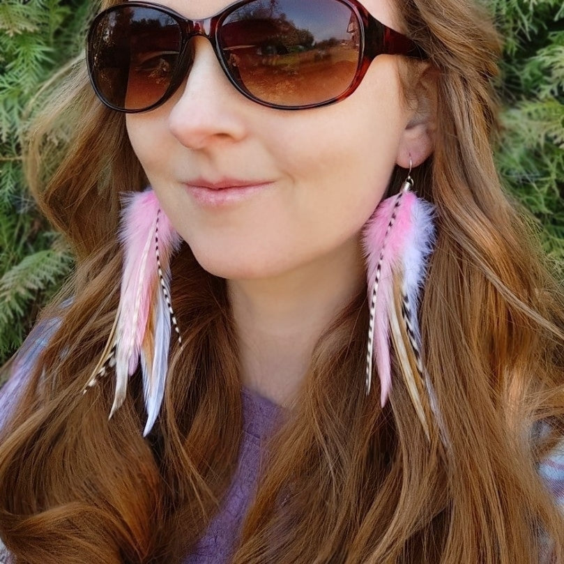 A model wearing long stripy feather earrings in pastel pinks and white. She is outside with greenery behind her.