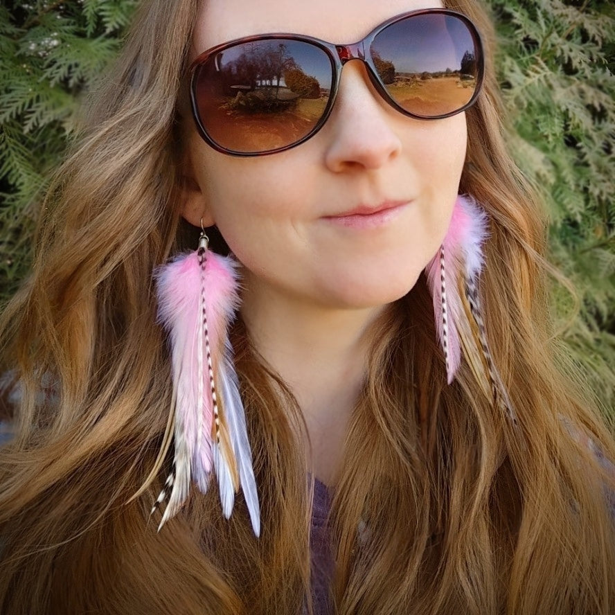 A model wearing long stripy feather earrings in pastel pinks and white. She is outside with greenery behind her.