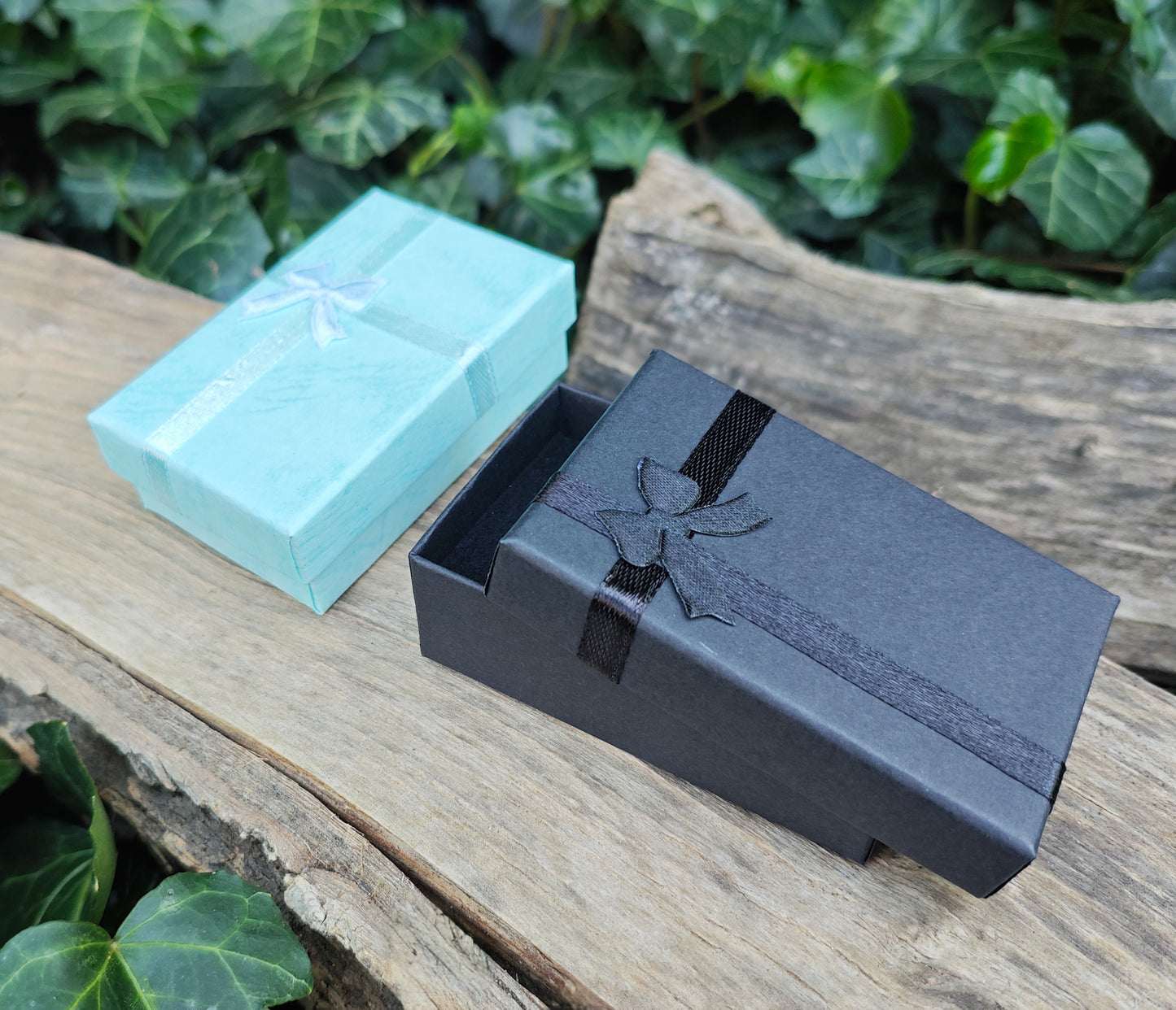 Two gift boxes, one black and one turquoise, on a wooden surface with green foliage in the background.