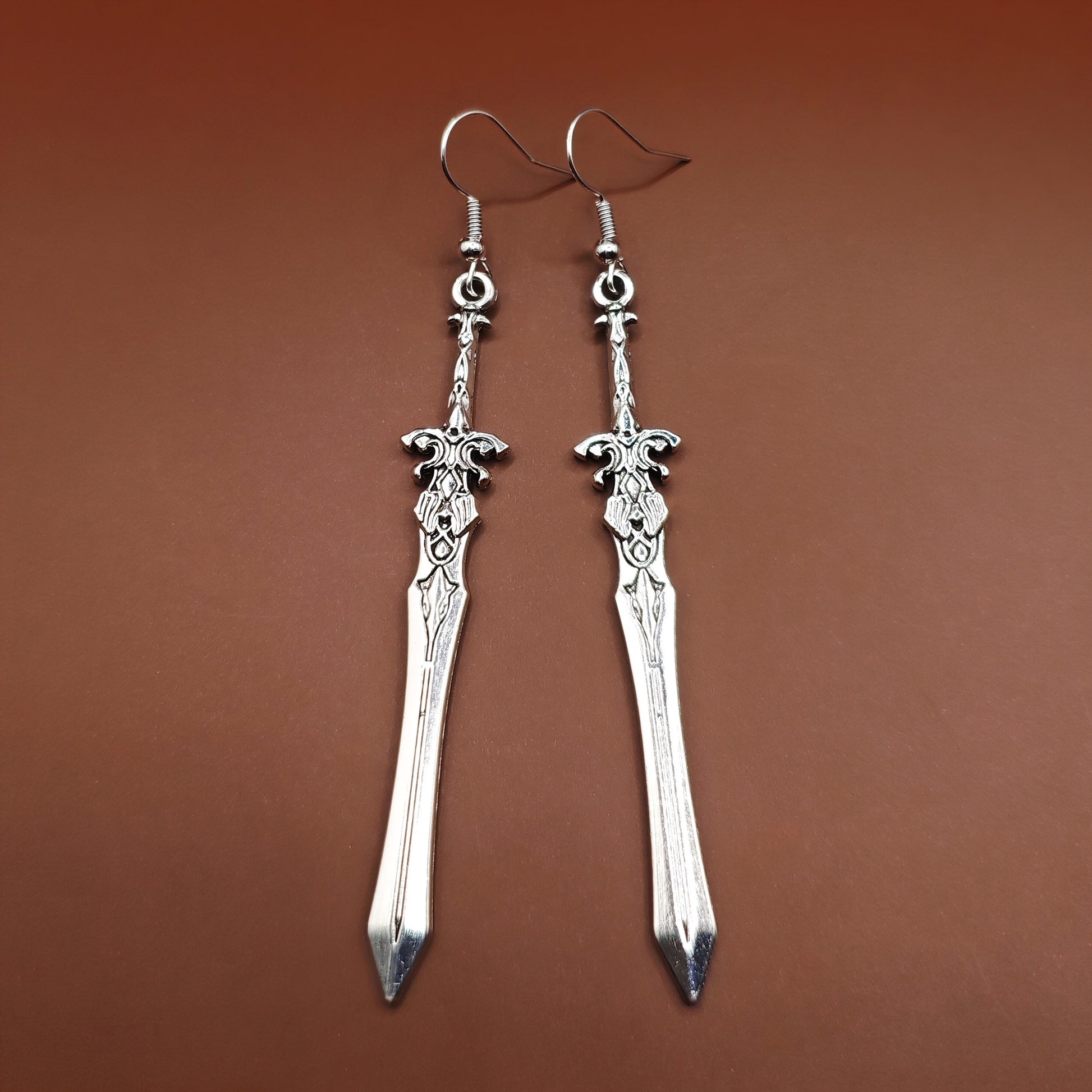 Medieval-style silver sword earrings dangling from stainless steel hooks on a brown background, perfect for fantasy fashion and cosplay.