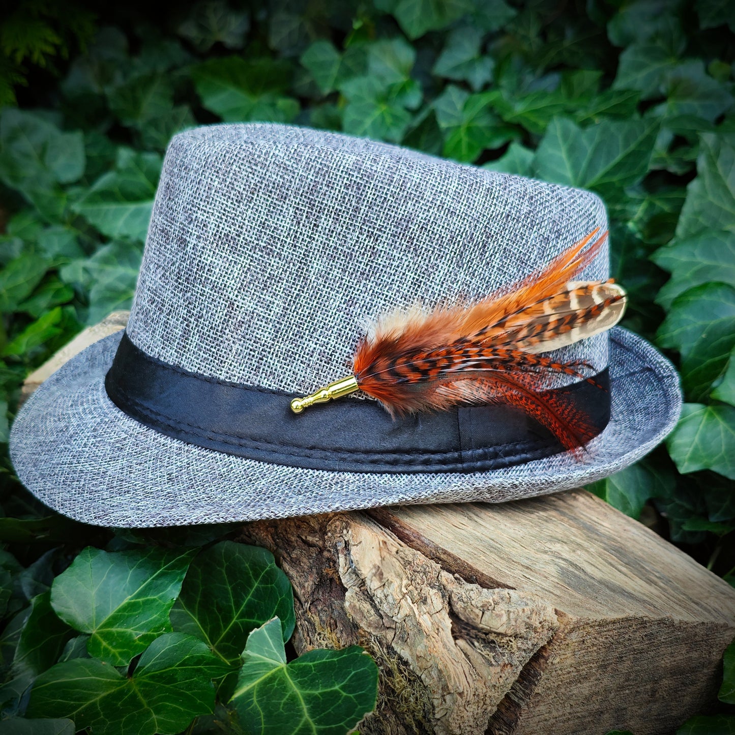 A funky red brown, orange and beige feather hat pin with gold pin on a light grey fedora hat. Modern hippie style. Green leaves in the background.