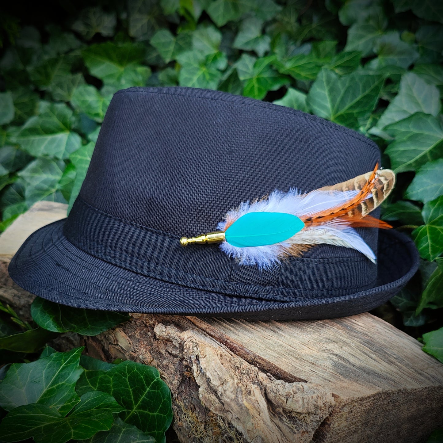 A colorful bright turquoise, orange, white and beige stripy feather hat pin with gold pin on a black bowler hat. Bold statement accessory. Green leaves in the background.