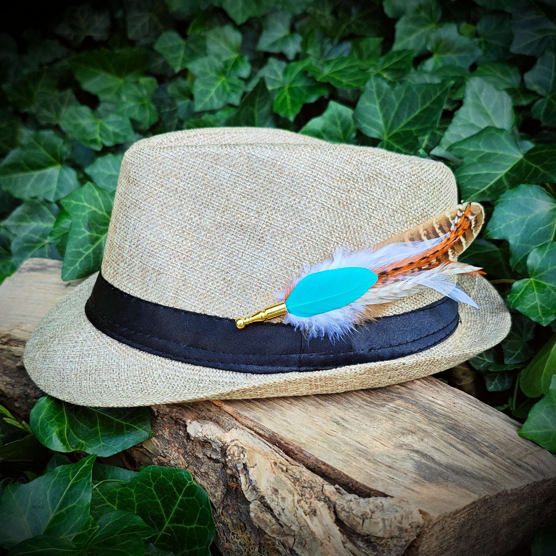 A stripy bright turquoise, orange, white and beige feather hat pin with gold pin on a light cream straw fedora hat. Modern hippie and boho style. Green leaves in the background.