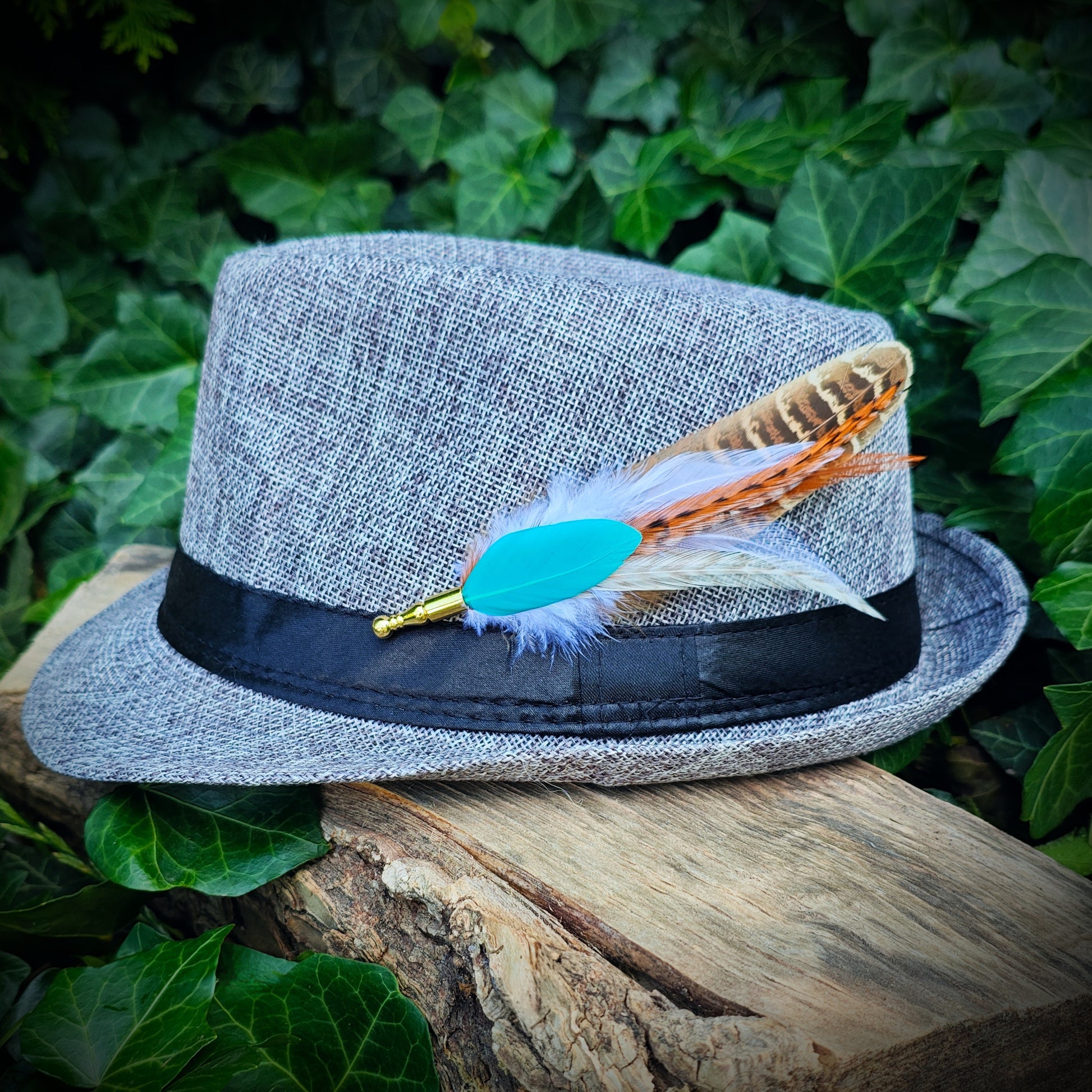 A stripy bright turquoise, orange, white and beige feather hat pin with gold pin on a light grey trilby fedora hat. Cute hippy style. Green leaves in the background.