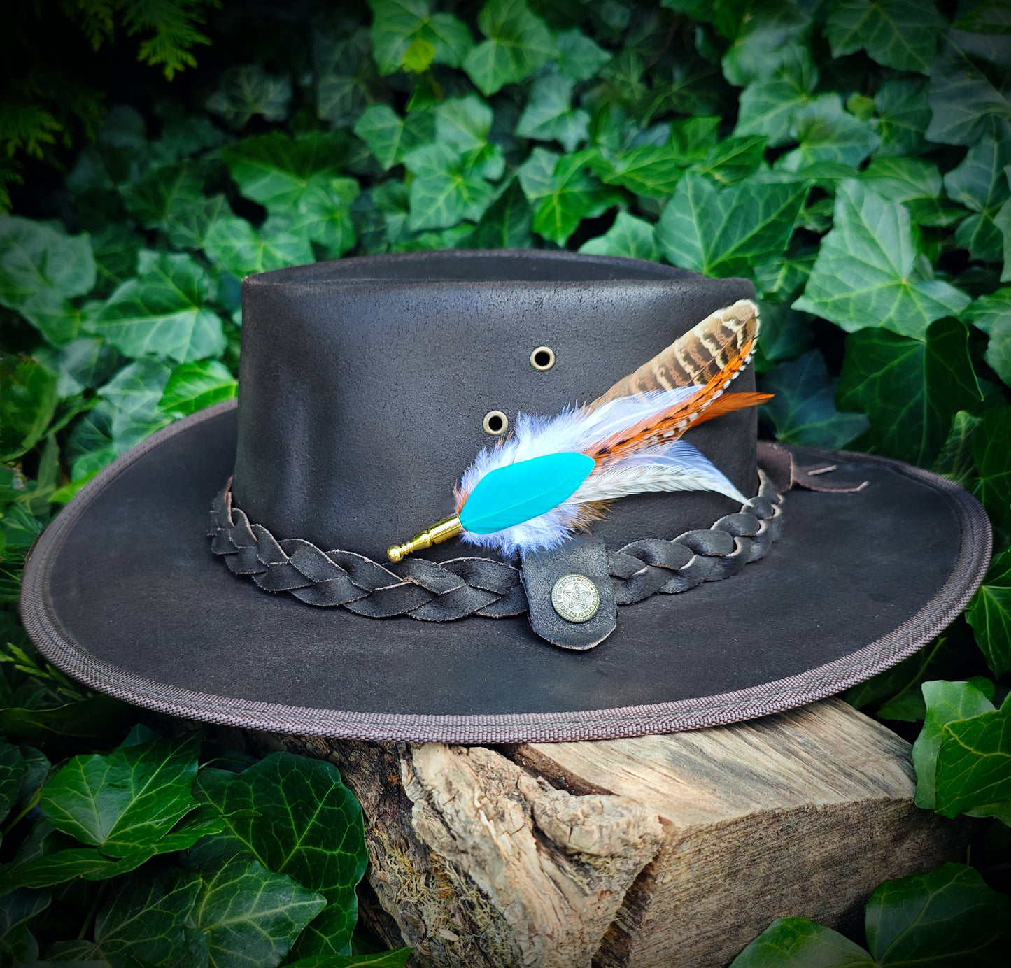 A stripy bright turquoise, orange, white and beige feather hat pin with gold pin on a brown leather cowboy hat. Cute western cowgirl style. Green leaves in the background.