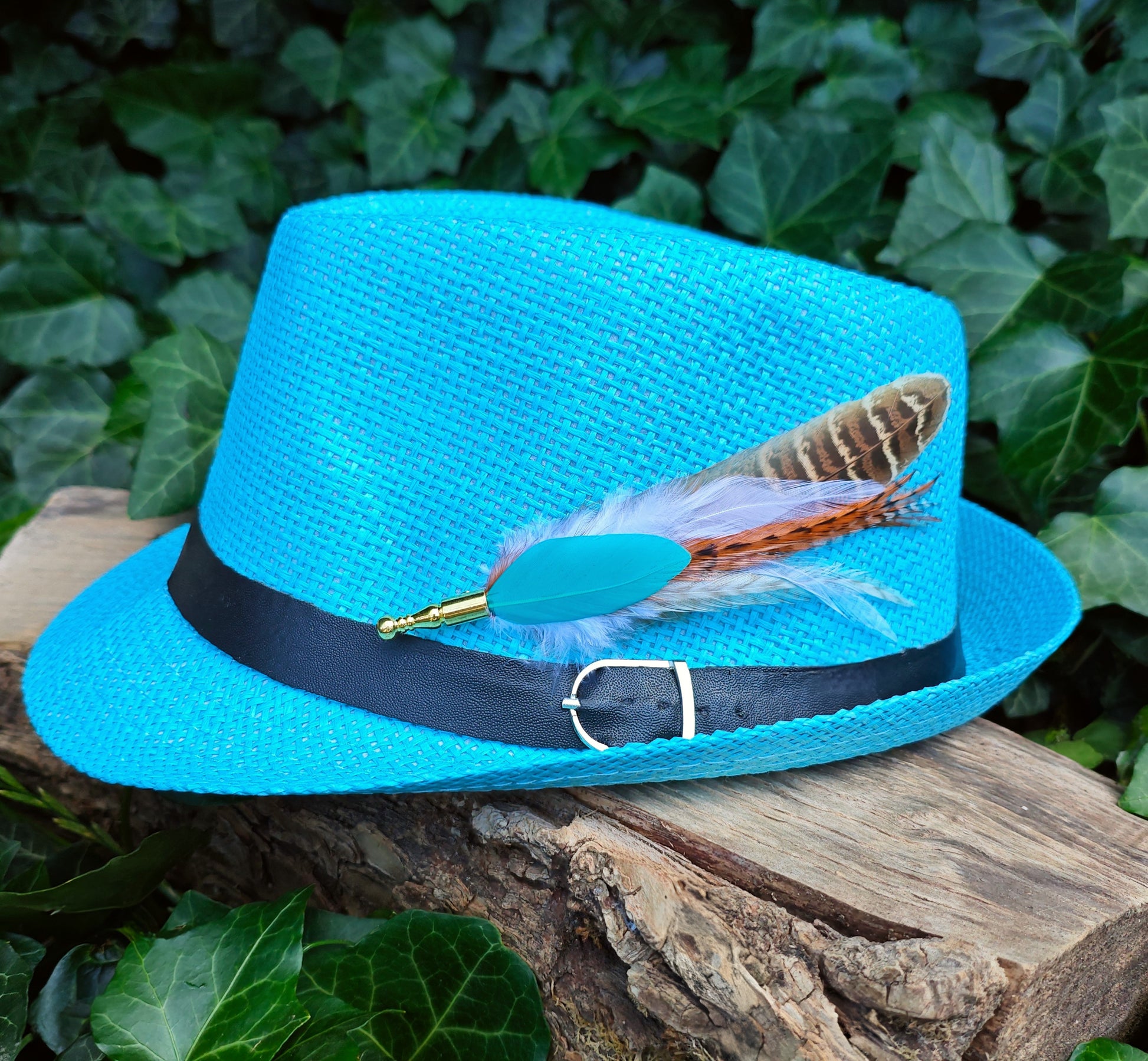A stripy bright turquoise, orange, white and beige feather hat pin with gold pin on a bright blue fedora hat. Bold funky style. Green leaves in the background.