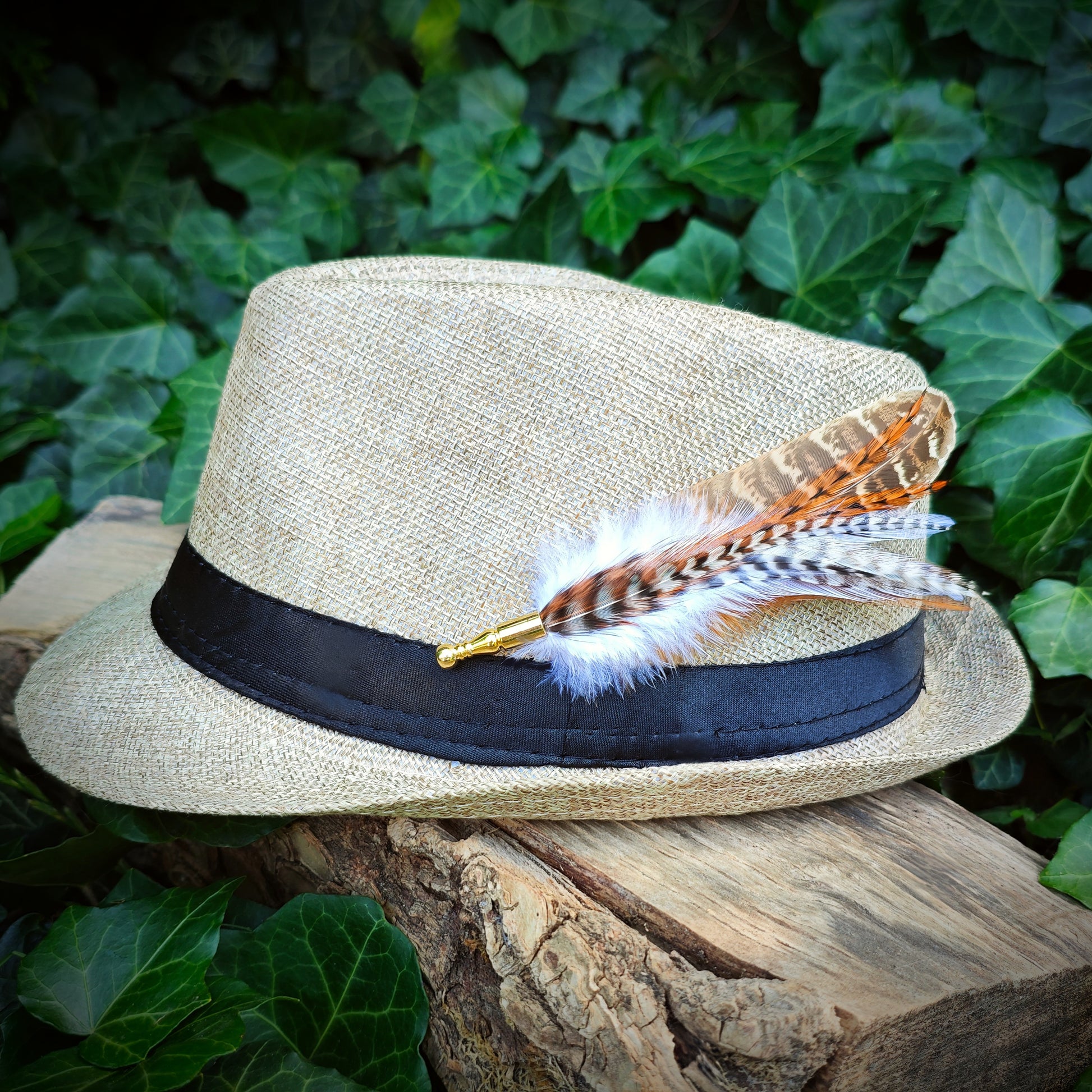 A brown and white feather hat pin, with a gold pin, on a beige straw hat on a wooden log with green foliage in the background