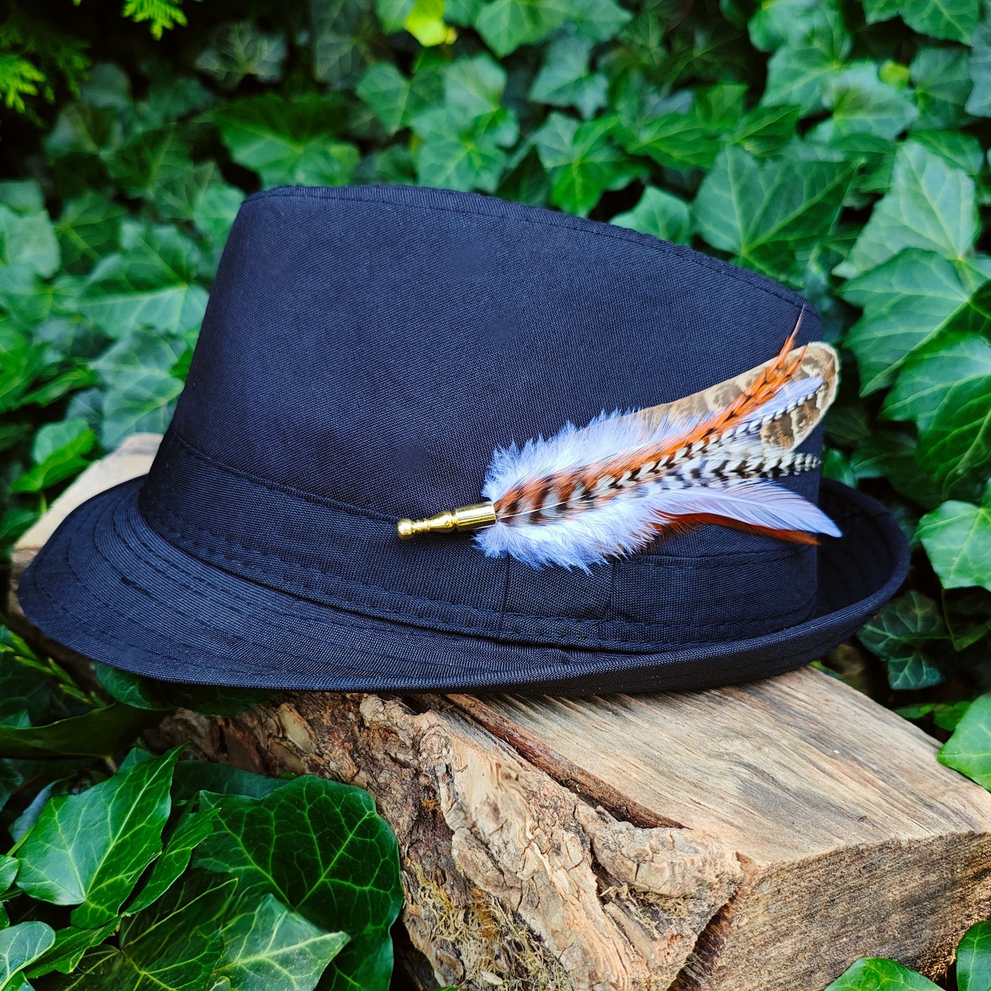 A brown, white and orange funky feather hat pin on a black bowler hat with greenery behind