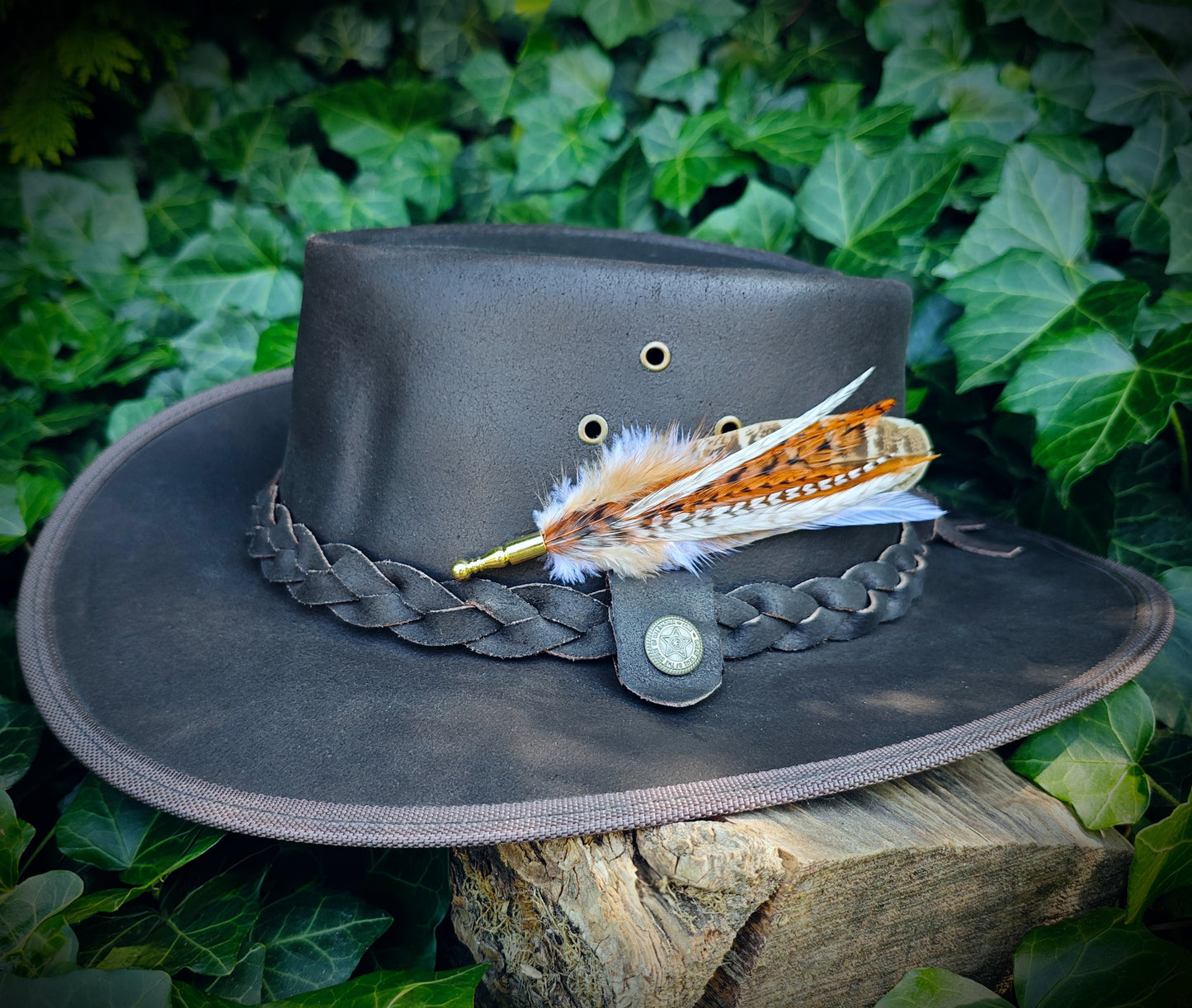 A stripy brown, orange and white feather hat pin on a brown leather cowboy hat with a gold pin. Western boho style. Green leaves in the background.