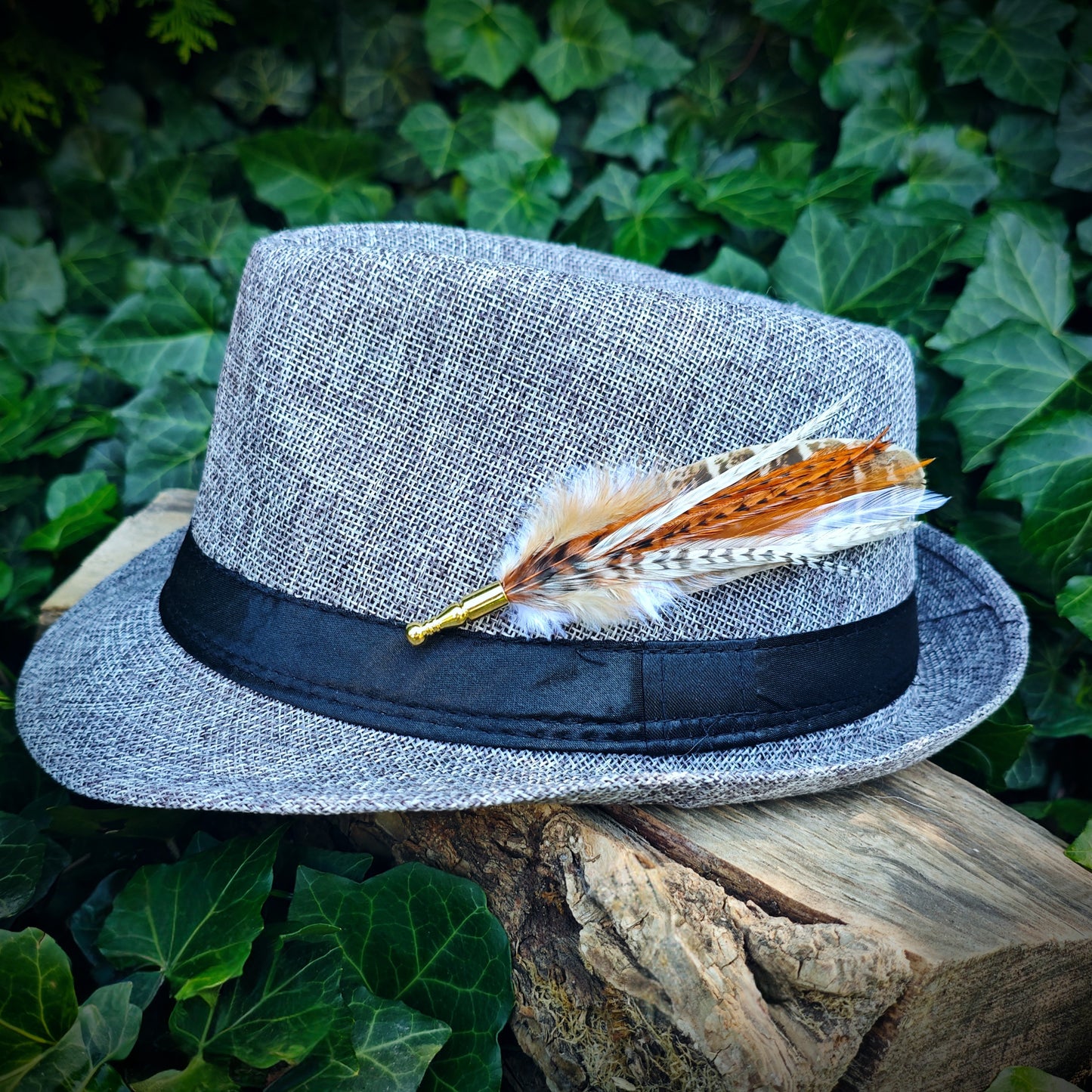 A rustic stripy brown, orange and white feather hat pin on a light grey fedora trillby hat with a gold pin. Hippie style. Green leaves in the background.