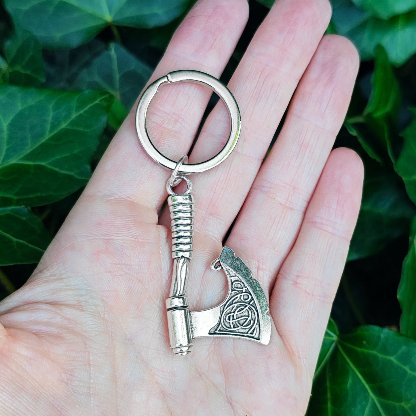 Silver medieval axe keychain held in a hand against a green leafy background