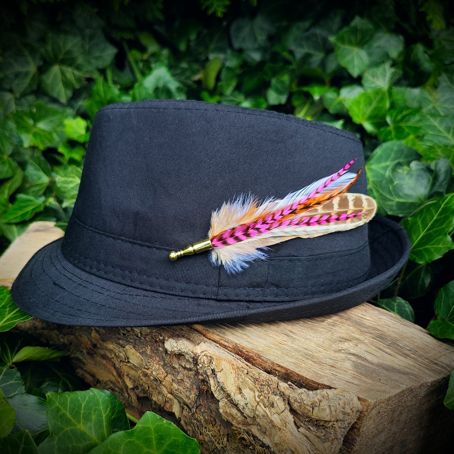 A stripy bright pink, orange, white and beige feather hat pin with gold pin on a black bowler fedora hat. Boho style. Green leaves in the background.