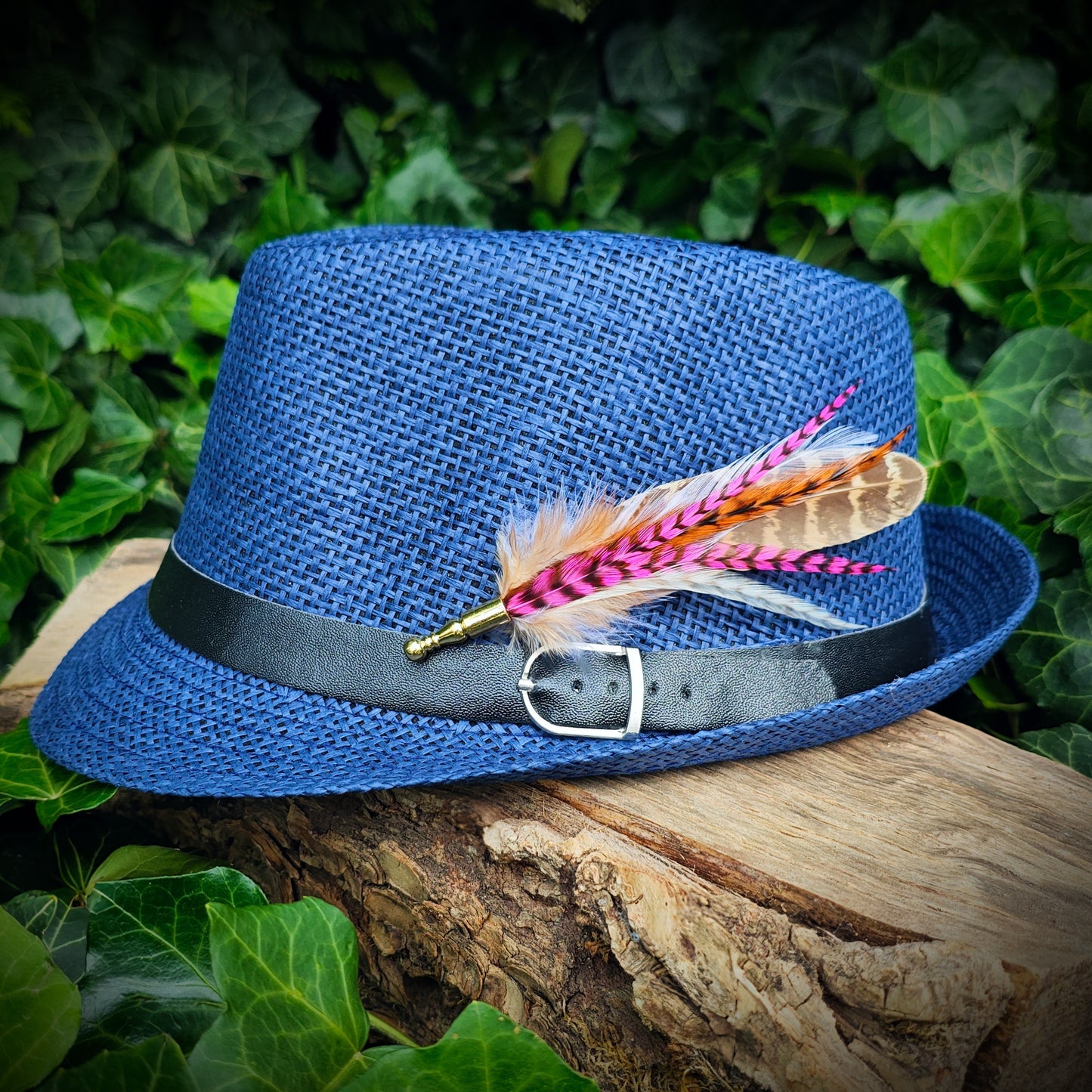A stripy bright pink, orange, white and beige feather hat pin with gold pin on a blue trilby fedora hat. Cute hippy style. Green leaves in the background.