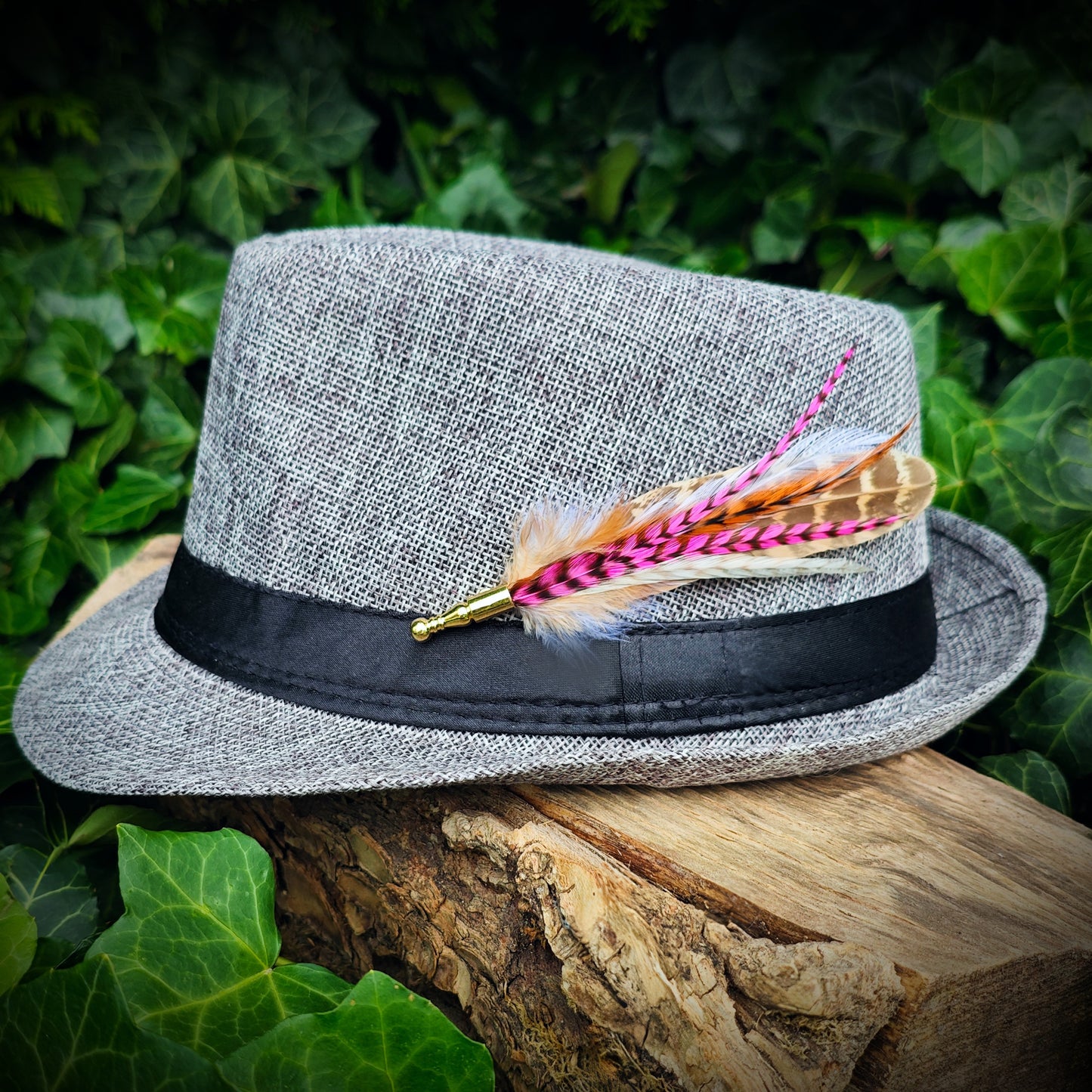 A stripy hot pink, orange, white and beige feather hat pin with gold pin on a light grey trilby fedora hat. Modern hippie style. Green leaves in the background.