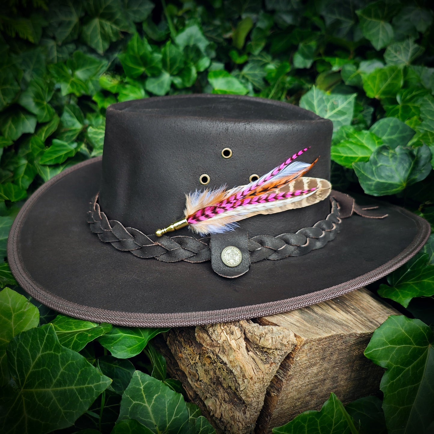 A stripy hot pink, orange, white and beige feather hat pin with gold pin on a brown leather cowboy hat. Vintage western cowgirl style. Green leaves in the background.