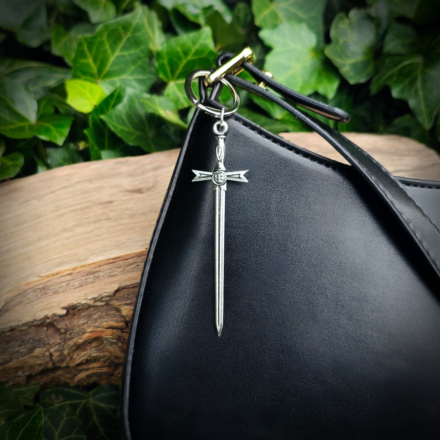 Black handbag with a silver sword keychain as a bag charm, against a green leafy background