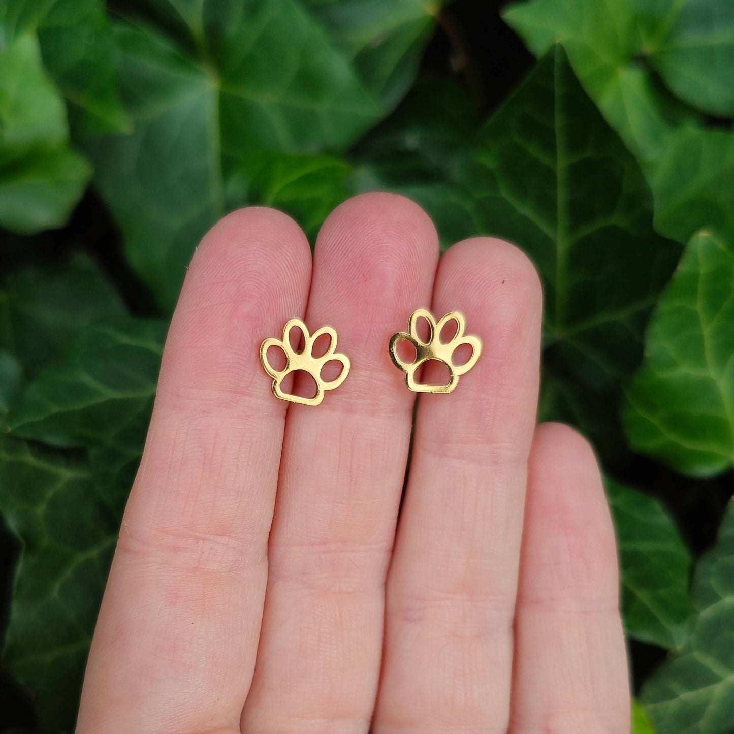 Gold paw print earrings held between fingers with a green leafy background. Gift for her.