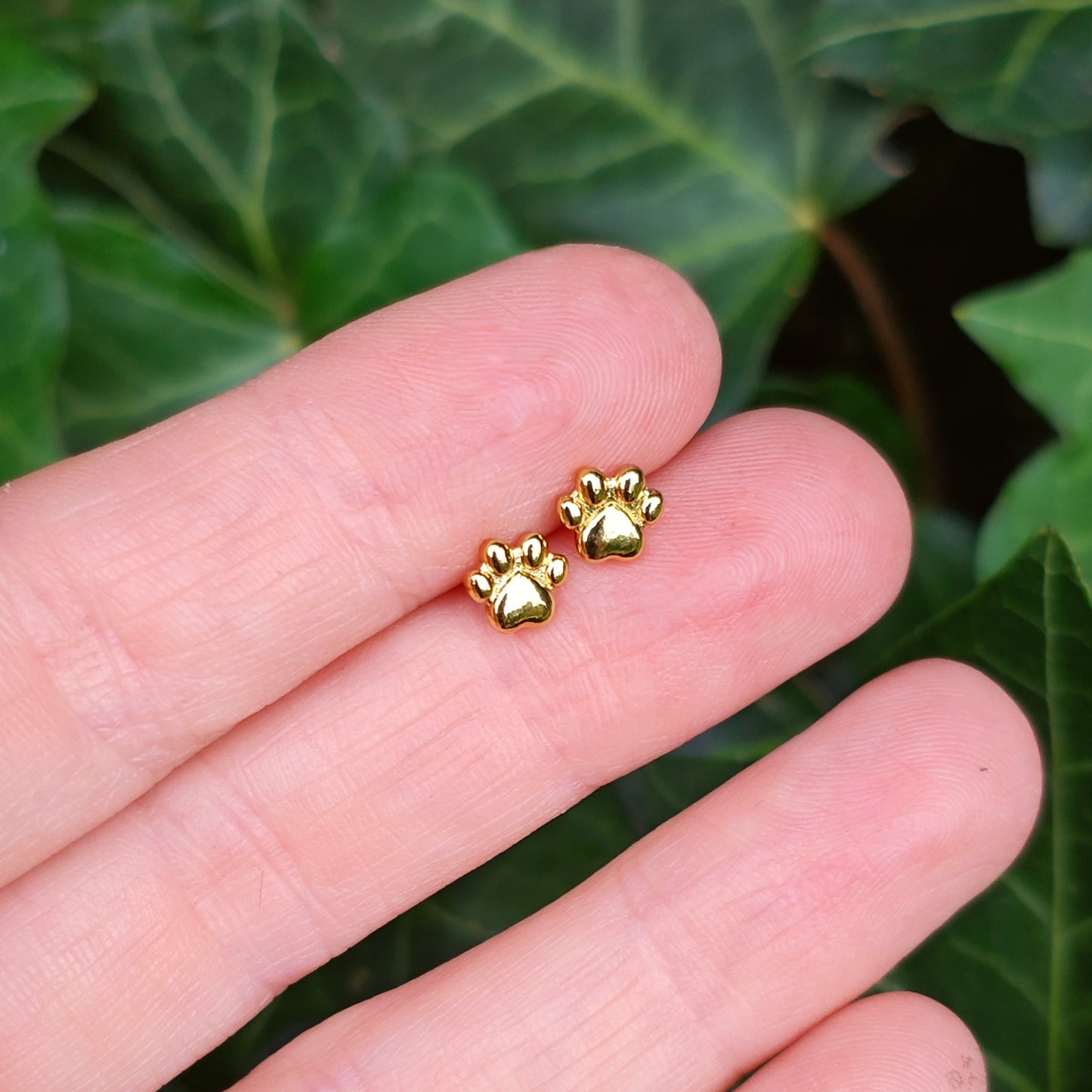 Gold paw-shaped earrings held between fingers with a green leafy background. A great gift idea for girls or animal lovers.