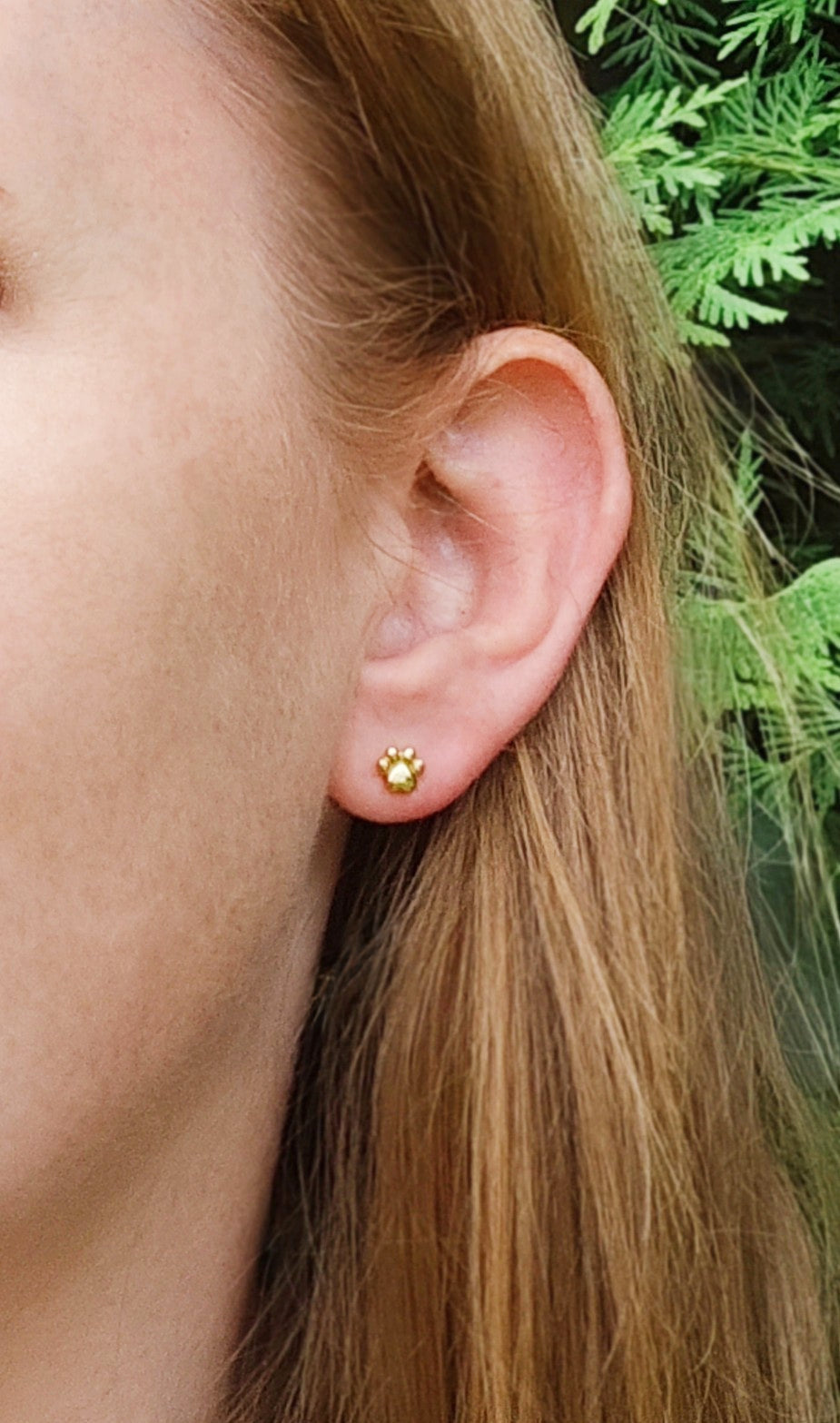 Close-up of an ear wearing a gold paw stud earring with a small design, against a green natural background.