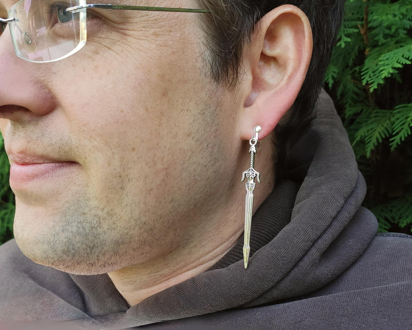 A male model wearing long sword earrings with silver hooks. He is outside, and looks cool, rugged and happy.