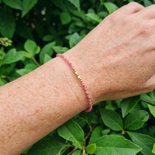 A wrist wearing a dainty boho beaded bracelet with natural pink strawberry quartz gemstone beads, outside with green leaves behind.