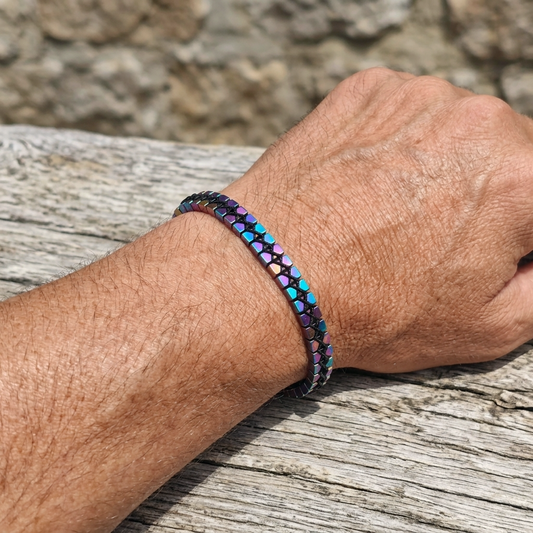 A man's wrist wearing a metallic multicolour rainbow Hematite beaded bracelet with cube beads and adjustable cord on a wooden surface