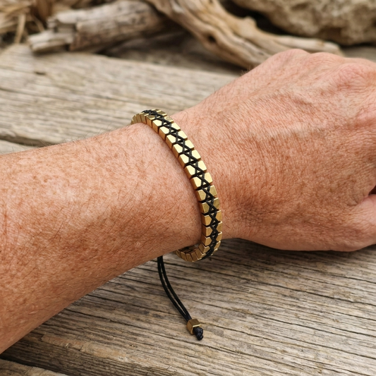 A man's wrist wearing a metallic gold Hematite beaded bracelet woven with cube beads and adjustable cord