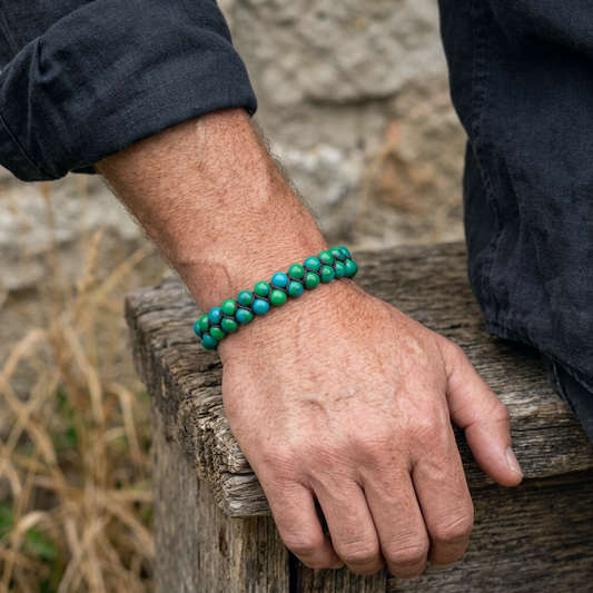 A man wearing a Chrysocolla bead bracelet featuring a double row of green and blue beads woven together. He sits on a wooden bench with a stone wall behind