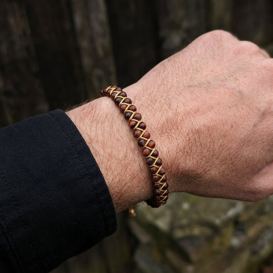 Close-up of a man's wrist wearing a braided red tigers eye bracelet with a blurred natural background.