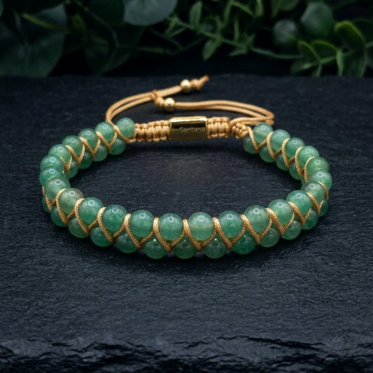 Green aventurine beaded gemstone bracelet with gold cord on a dark stone surface with green leaves in the background