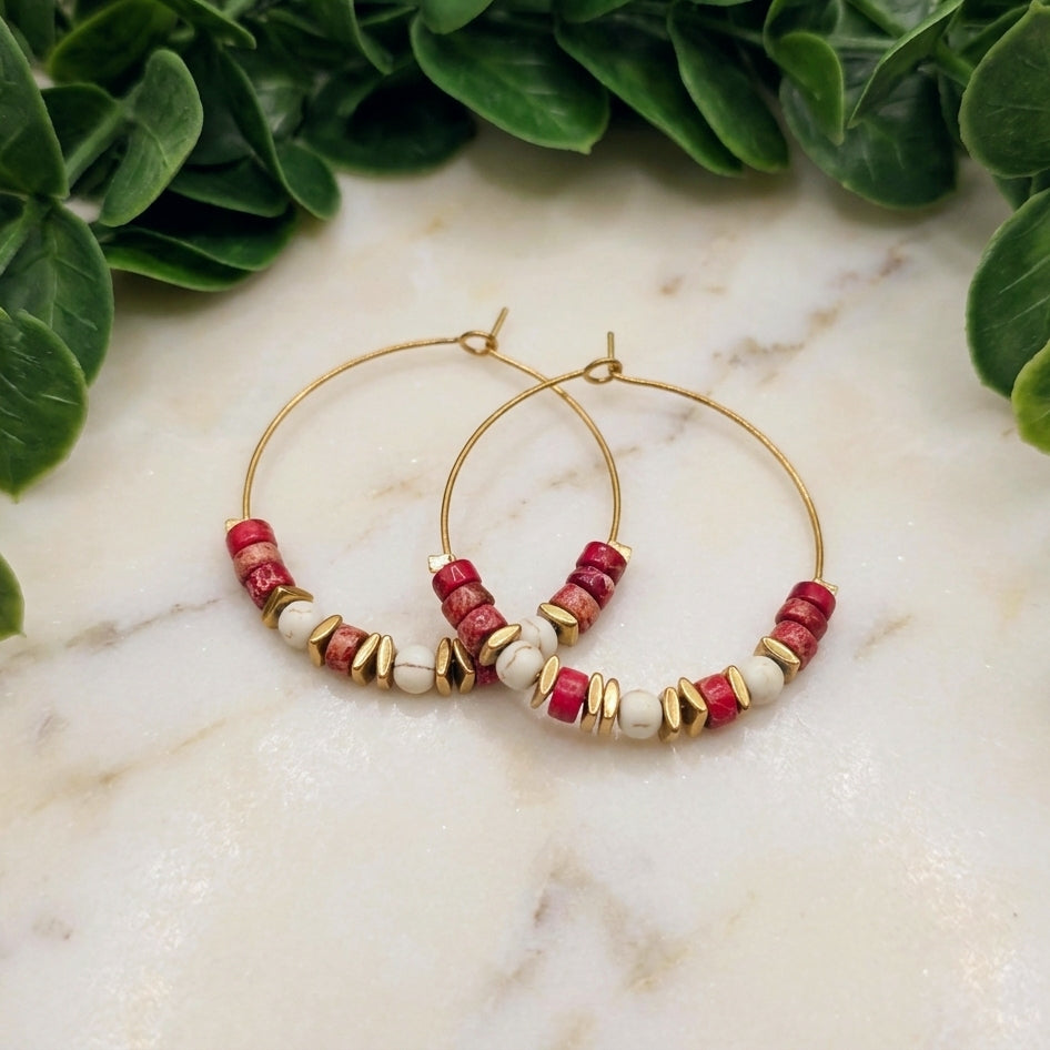 Gold hoop earrings with red and white marbled beads, displayed on a white marble surface with green leaves in the background.