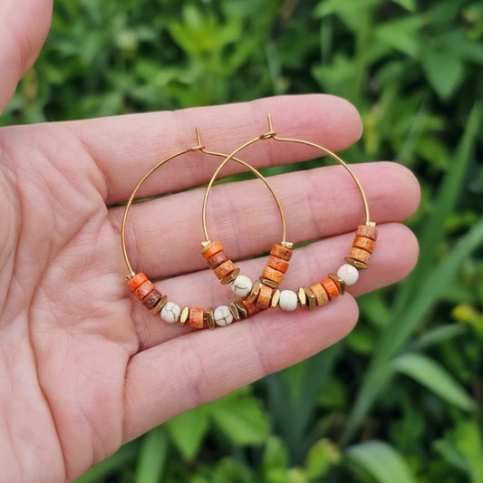 A pair of gold hoop earrings with small orange jasper and white beads, held in a person's hand against a blurred green outdoor background.
