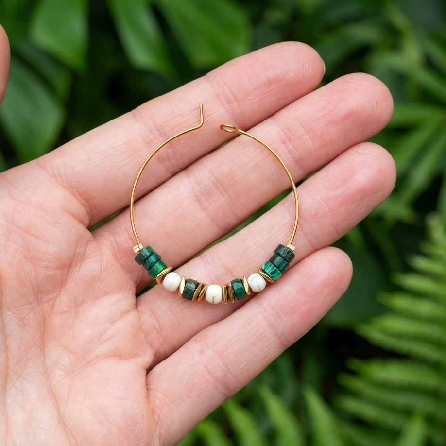 A gold hoop earring with green and white beads, held in a person's hand against a blurred background of green foliage.