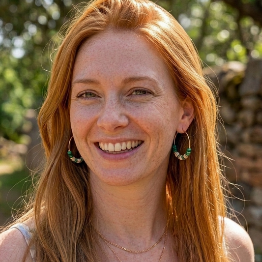A smiling woman with long red hair wearing green and white beaded hoop earrings, photographed outdoors with a blurred natural background.