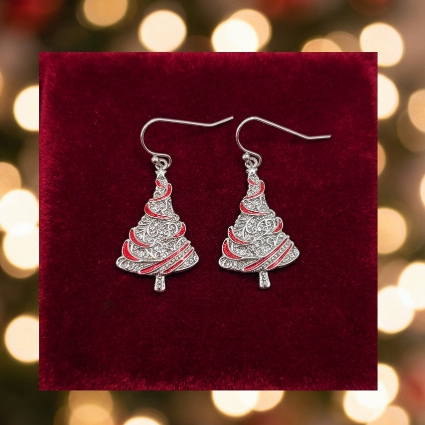 A pair of silver-toned dangle earrings shaped like Christmas trees with red enamel stripes, displayed on a dark red velvet surface against a blurred holiday light background.