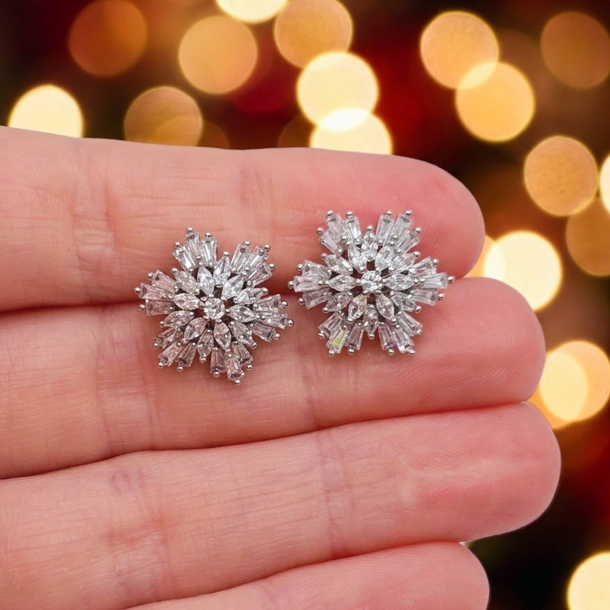 A pair of snowflake-shaped stud earrings with clear baguette-cut stones in a silver-toned setting, displayed on a person's fingers against a warm, blurred holiday bokeh background.