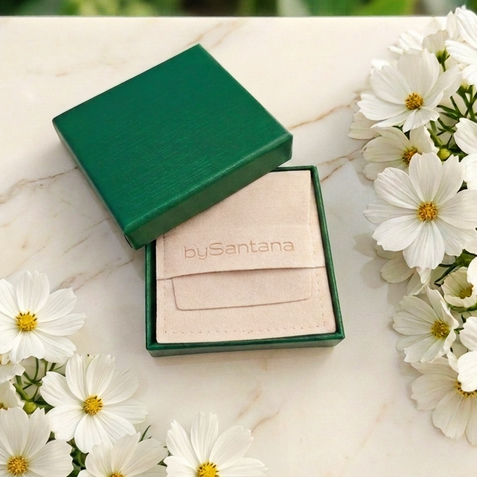 A green jewelry gift box with its lid open, revealing a beige velvet pouch embossed with the brand name 'bySantana'. The box is styled on a white marble surface and surrounded by white daisies.