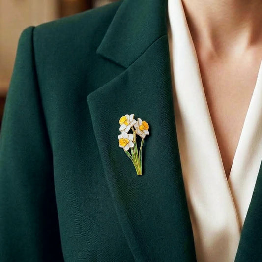 A close-up of a person wearing a dark green blazer over a cream satin V-neck top, featuring a small enamel daffodil brooch pinned to the lapel.