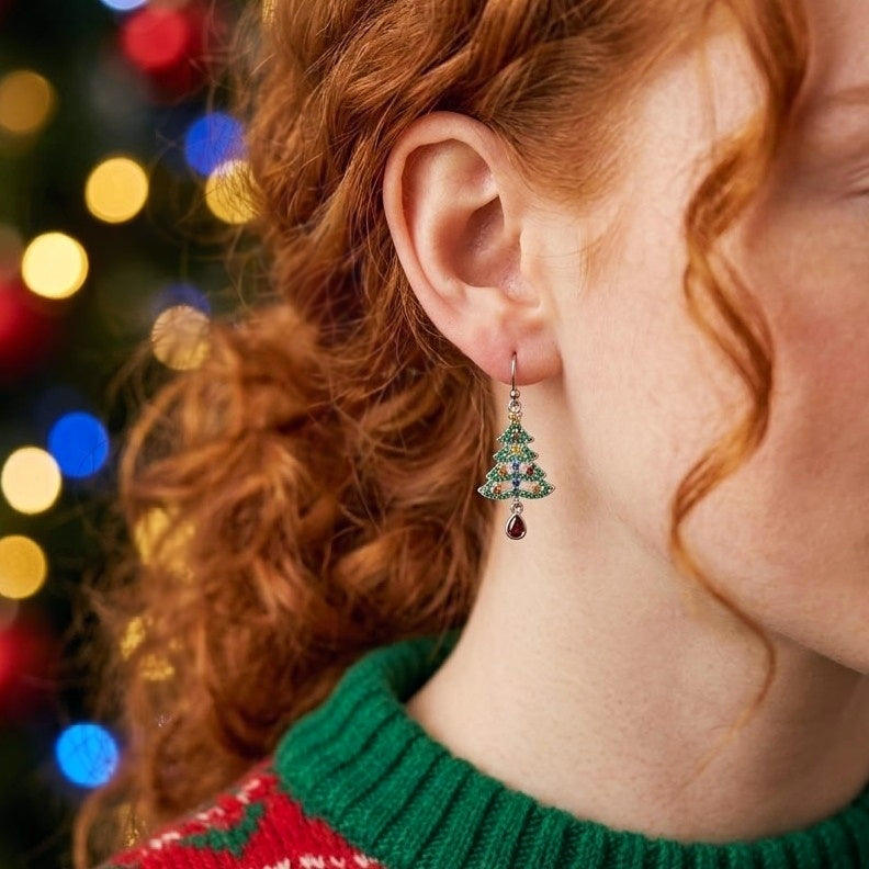 A close-up of a person with red hair wearing a dangling Christmas tree earring. The earring features green and blue cubic zirconia crystals with a small dark red teardrop charm.
