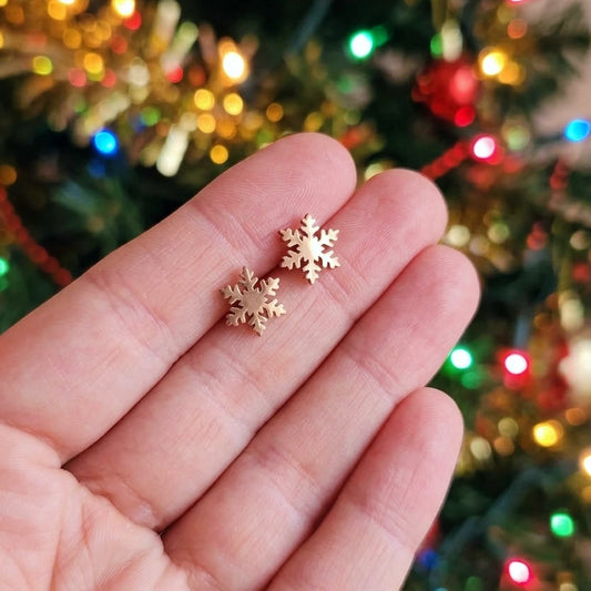 Two small rose gold snowflake stud earrings held on a person's fingers, with a blurred Christmas tree and colorful lights in the background.