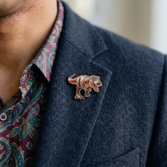 A close-up of a person wearing a small metallic Tyrannosaurus Rex dinosaur pin on a dark blazer over a paisley shirt.