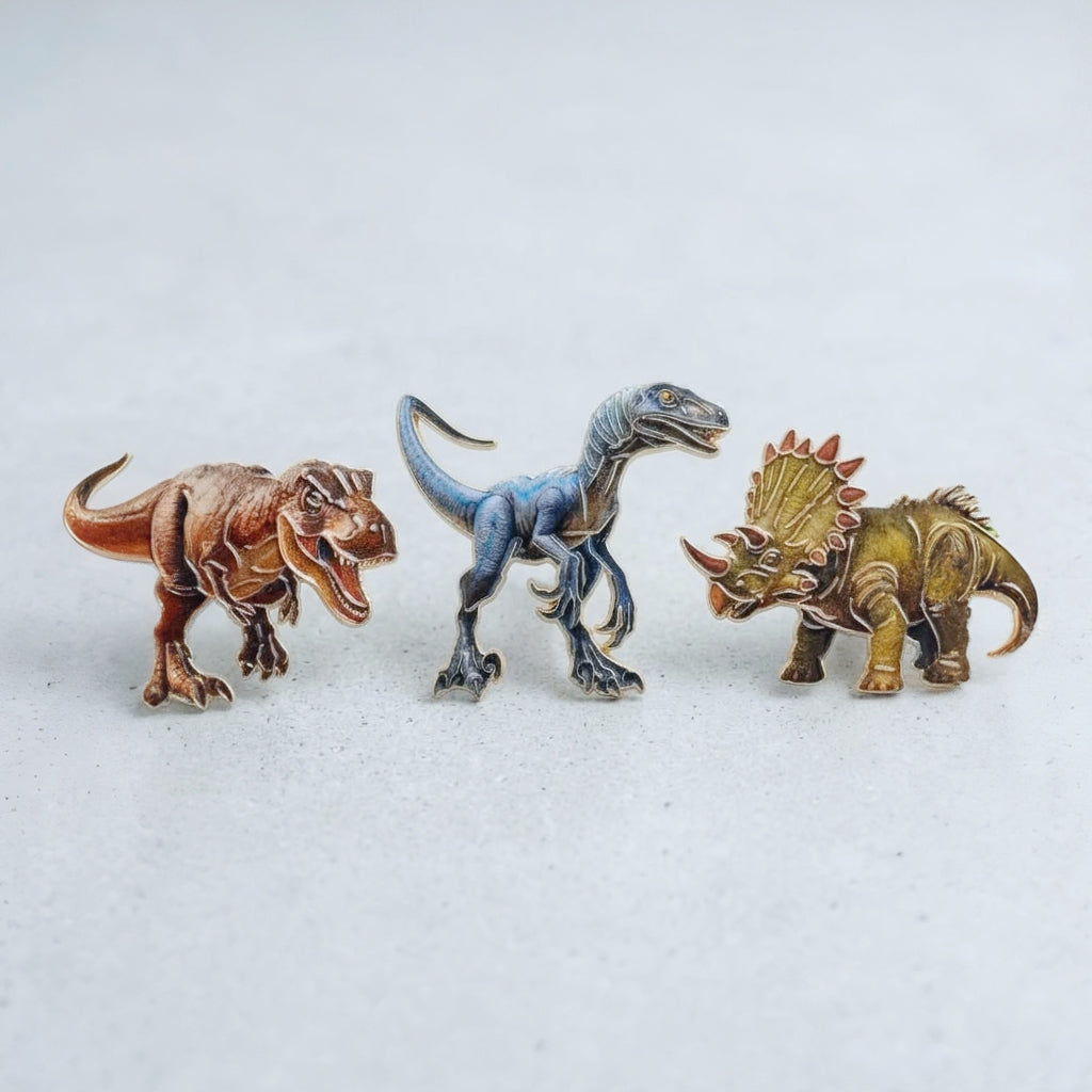 Three small dinosaur pins arranged side by side on a light gray surface: a brown t-rex on the left, a blue raptor dinosaur in the center, and a gold triceratops on the right.