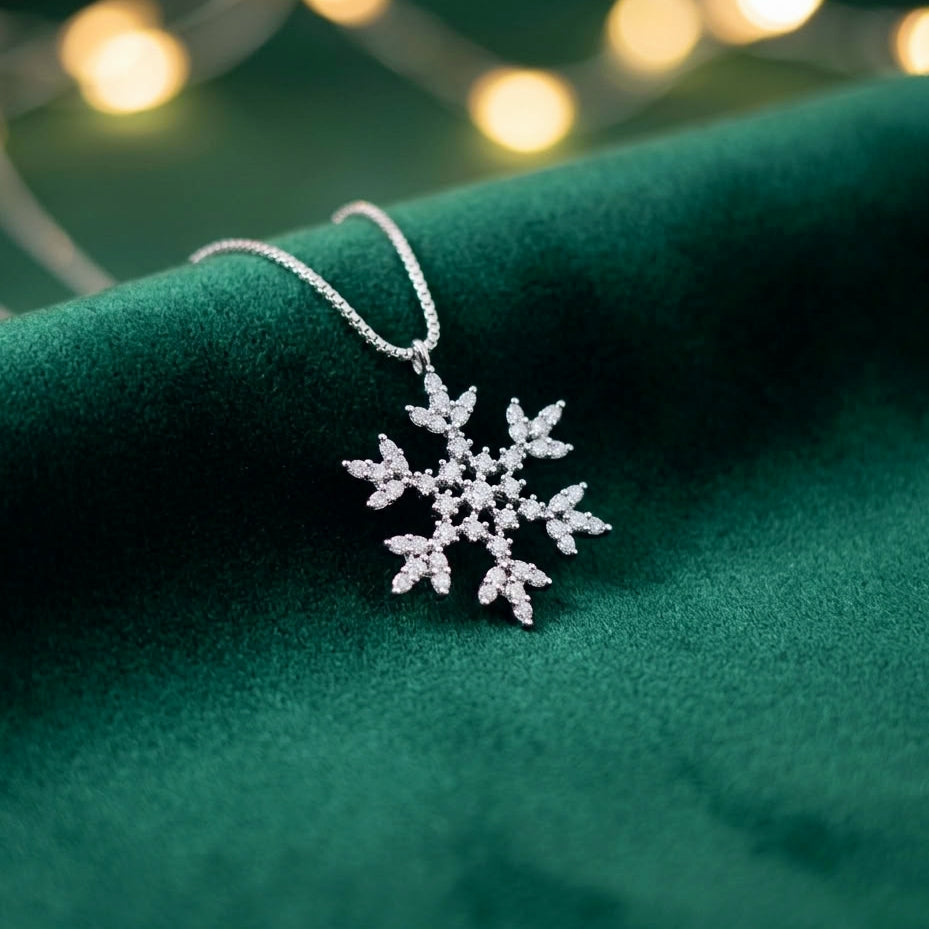 A silver snowflake pendant necklace adorned with small clear stones, resting on a dark green velvet fabric with blurred warm string lights in the background.