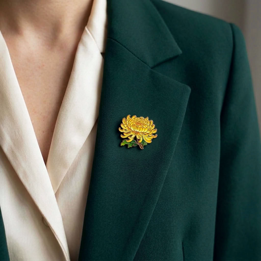 A close-up of a person wearing a dark green blazer over a cream satin shirt, featuring a yellow chrysanthemum and green floral enamel pin on the lapel.