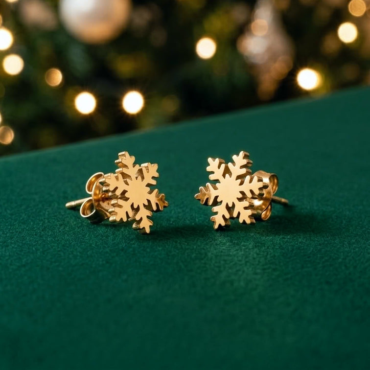 A pair of gold snowflake-shaped stud earrings displayed on a dark green velvet surface with a blurred Christmas tree and warm lights in the background.