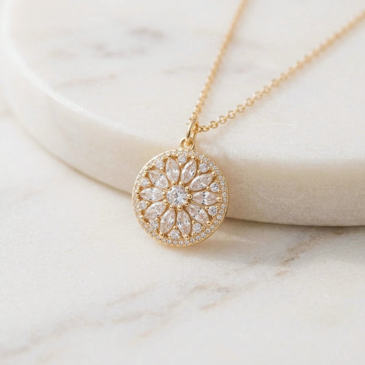 A gold circular pendant necklace with a floral sunflower sunburst design set with clear stones, resting on a white marble surface.