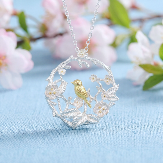 Silver necklace with a gold bird and flowers pendant on a light blue fabric background with pink flowers.