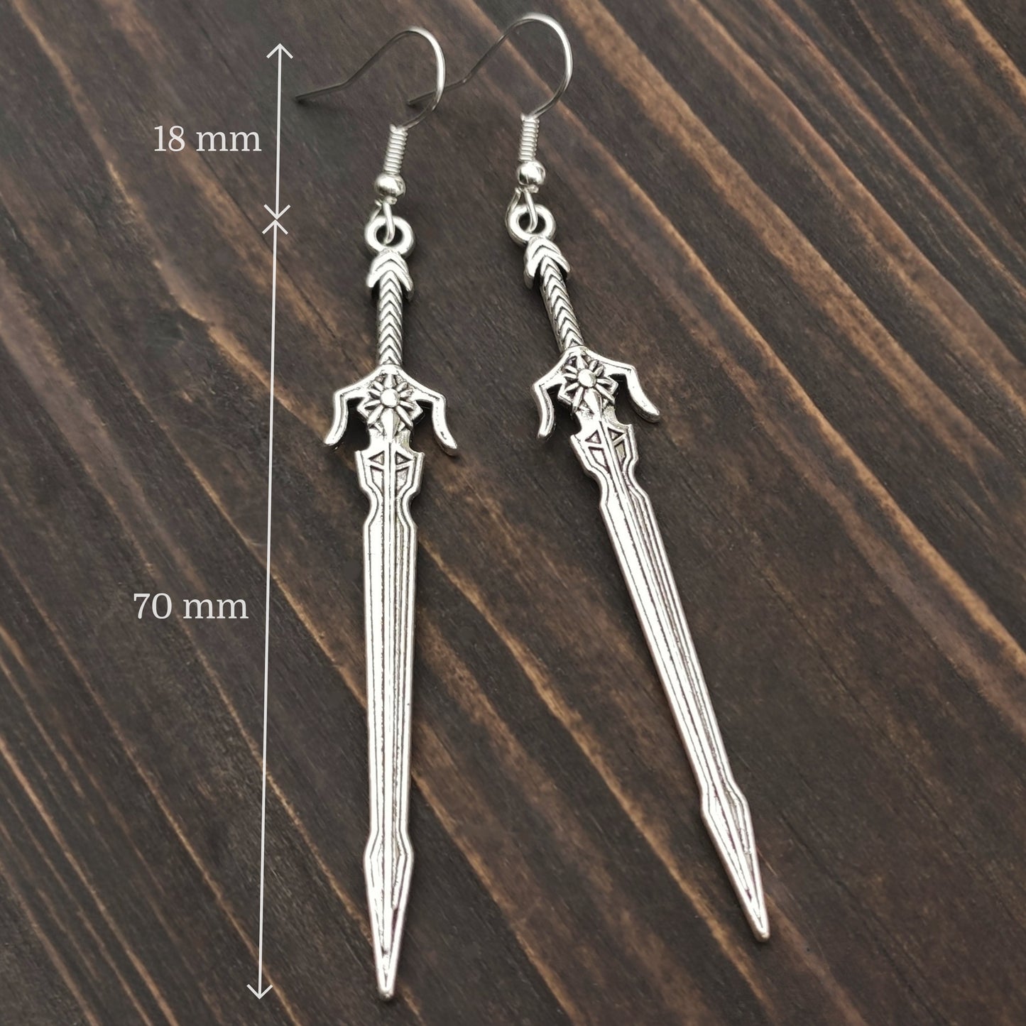 Silver sword earrings with hooks on a wooden surface with measurements indicated.